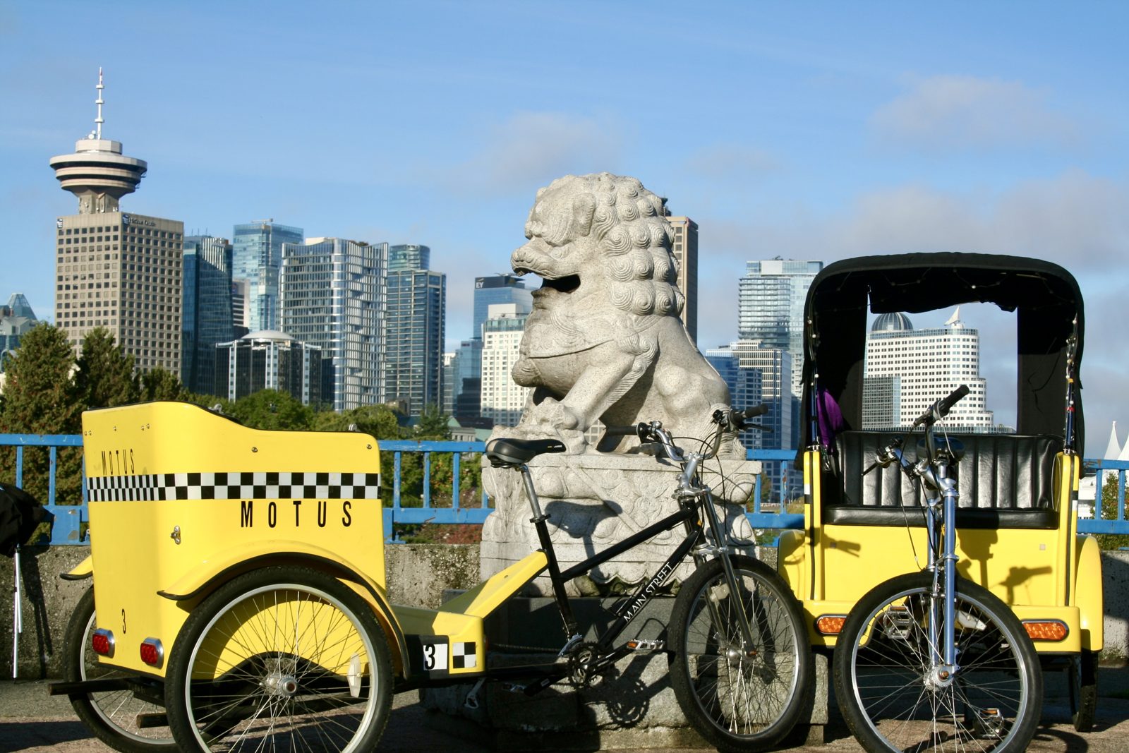 Two yellow Motus pedicabs beside a stone lion statue, with the downtown Vancouver skyline and Harbour Centre behind